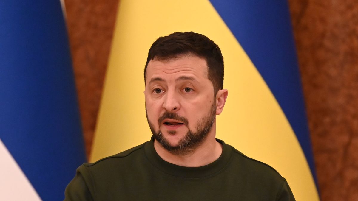 Prezydent Ukrainy Wołodymyr Zełenski w zielonym swetrze na tle flagi ukraińskiej w czasie konferencji prasowej