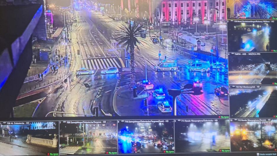 Policyjne radiowozy z włączonymi sygnałami świetlnymi blokują ruch na mokrej ulicy, w tle widoczne oświetlenie miejskie.