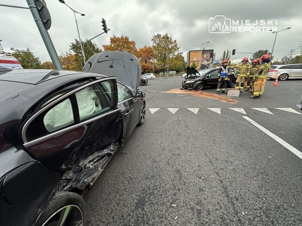 Czarny samochód osobowy z uszkodzoną boczną częścią stoi na skrzyżowaniu, obok pracują strażacy.