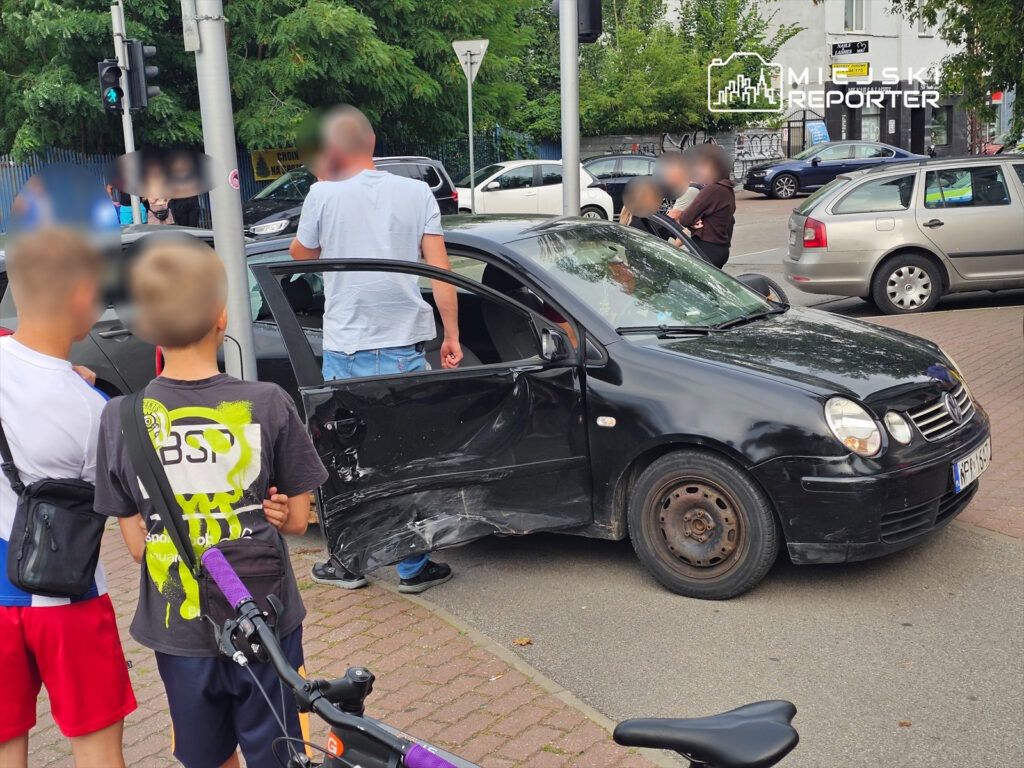 Mężczyzna rozmawia z innym mężczyzną obok uszkodzonego czarnego samochodu na ruchliwej ulicy, obok grupy dzieci.