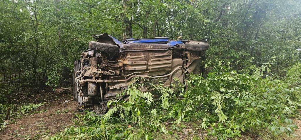 Wypadek samochodowy w lesie. Na zdjęciu przewrócone auto osobowe, które wpadło w zarośla. Widoczne uszkodzenia pojazdu oraz otaczająca go roślinność. Sytuacja wskazuje na niebezpieczne zdarzenie drogowe. Brak osób poszkodowanych w zasięgu kadru.