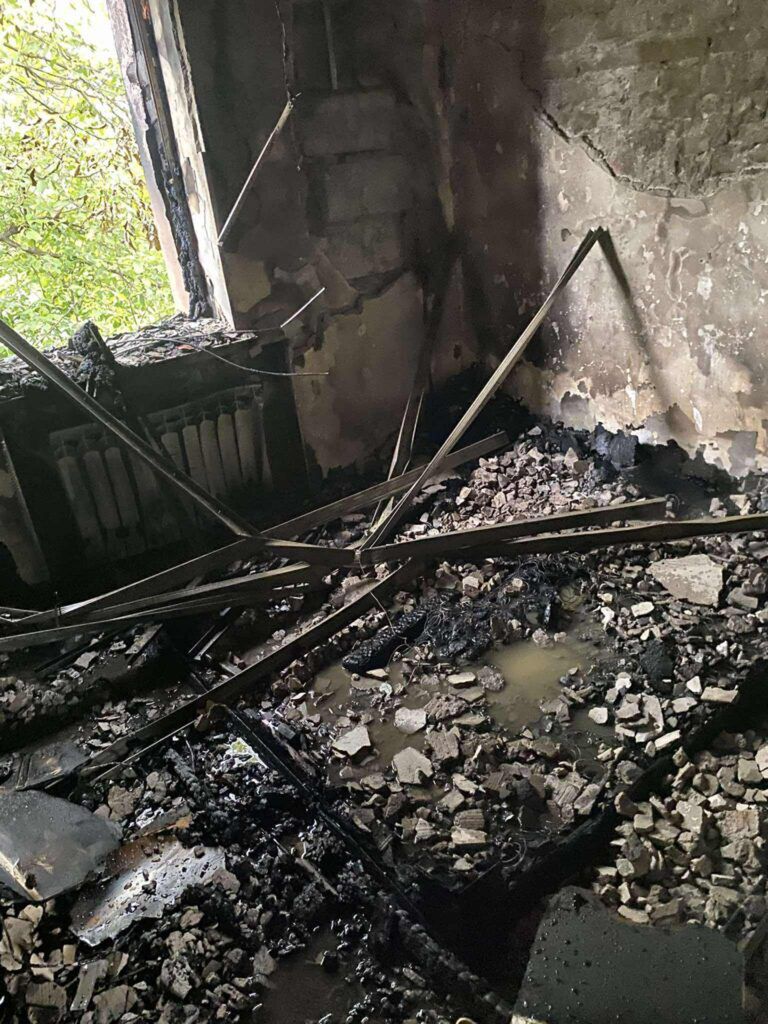 Zniszczony pokój z zawalonymi ścianami i podłogą pokrytą gruzem oraz wodą, z widocznymi śladami po pożarze.