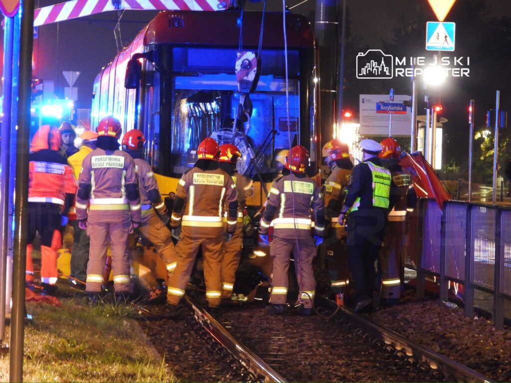 Grupa ratowników w kamizelkach odblaskowych pracuje przy tramwaju, który uległ wypadkowi, w nocy na torowisku.