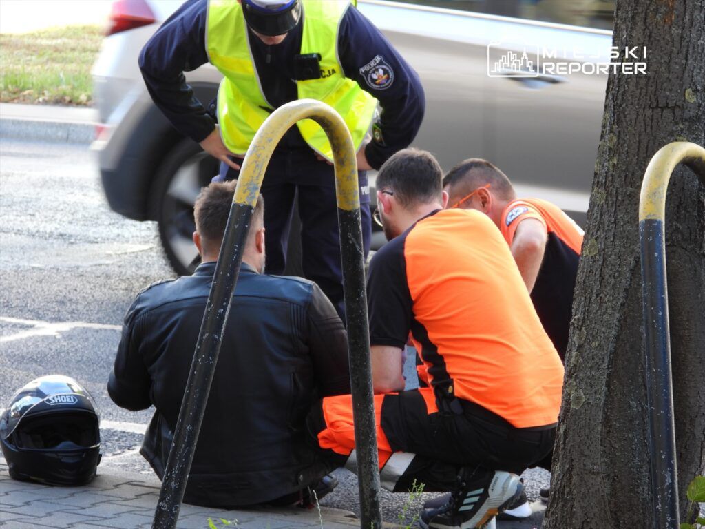 Czterej mężczyźni w odzieży roboczej otaczają osobę siedzącą na chodniku, obok leży kask motocyklowy.