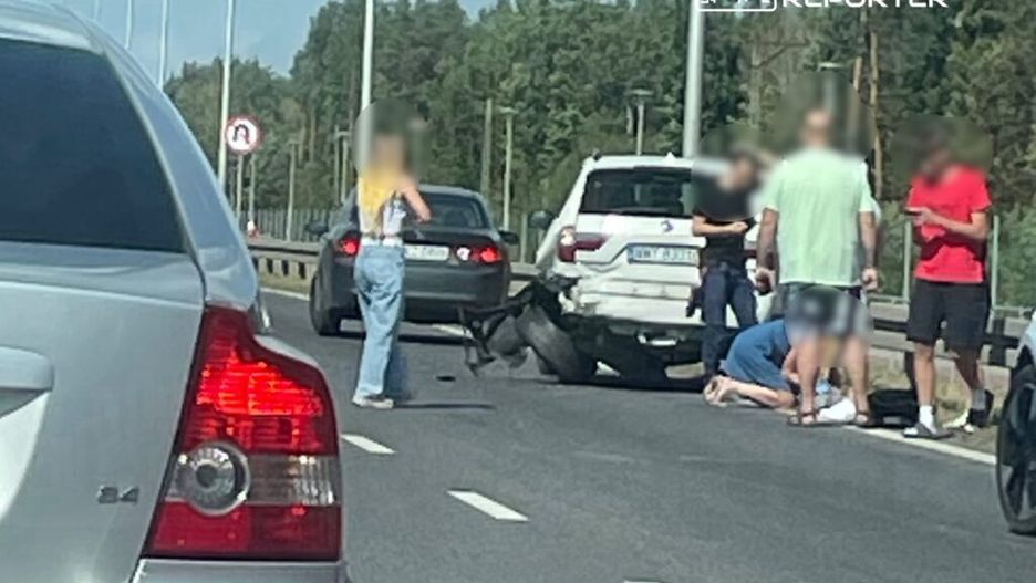 Na drodze stoi mężczyzna w krótkich spodniach, pomagający osobie leżącej na jezdni, obok grupka ludzi.