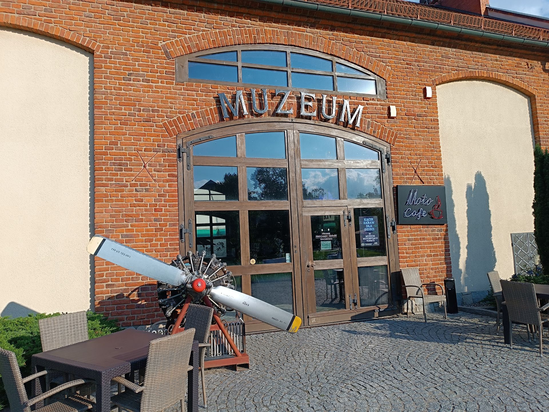 Ceglana fasada muzeum Zamek Topacz z napisem MUZEUM nad przeszklonym wejściem i eksponatem śmigła przed budynkiem