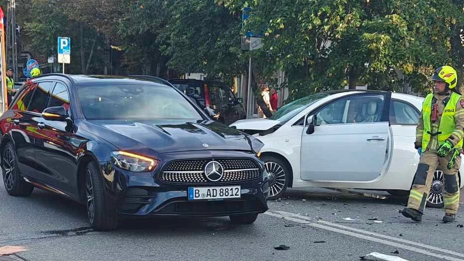 Czarny samochód osobowy z przodu, obok uszkodzony biały pojazd, a obok nich strażak w mundurze, zbierający szczątki.