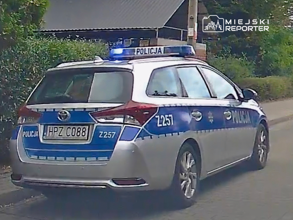 Policyjny radiowóz z niebieskimi światłami zaparkowany na ulicy, otoczony zielenią.