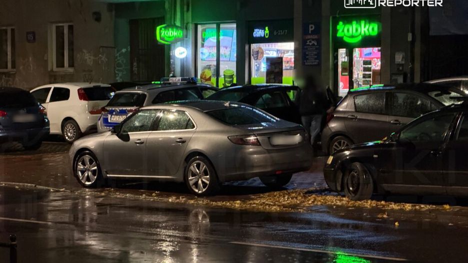 Srebrny samochód zaparkowany na mokrej ulicy, obok sklepu Żabka z podświetlonym znakiem. W tle inne samochody i policja.