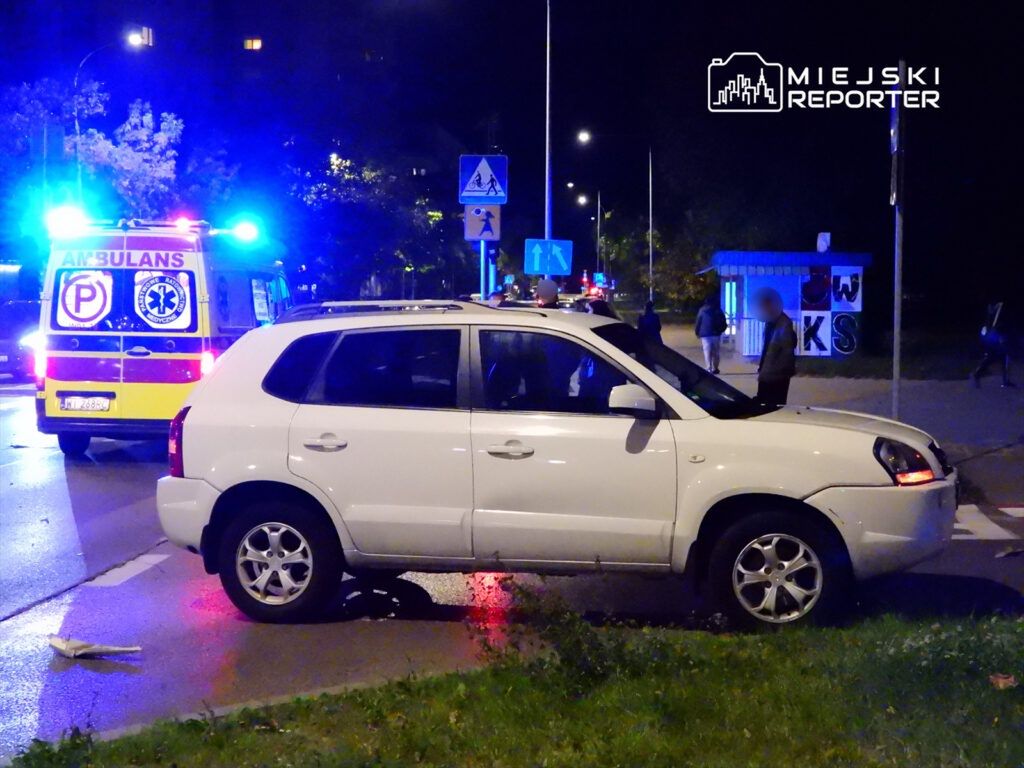 Biały samochód osobowy zatrzymany na skrzyżowaniu, w tle widać ambulans z włączonymi sygnałami świetlnymi.