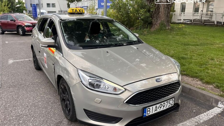 Taksi Ford stojące na parkingu, z widocznym znakiem "TAXI" na dachu i uszkodzeniami na boku.