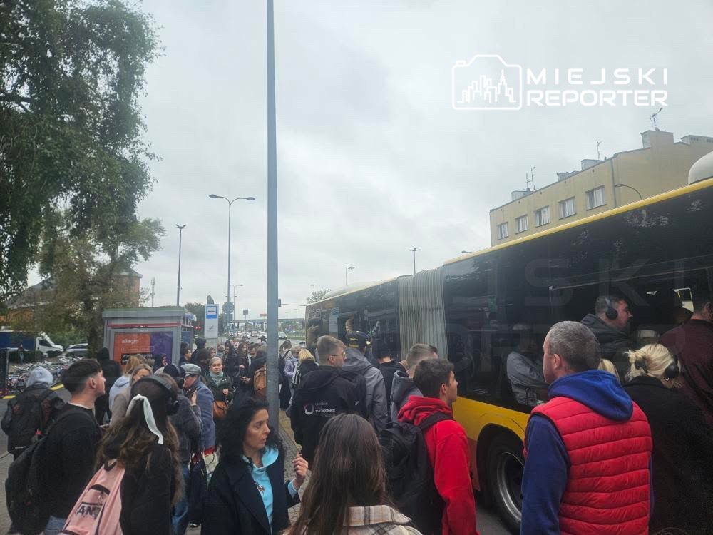 Tłum ludzi czeka na przystanku autobusowym w pobliżu dużego, żółtego autobusu.