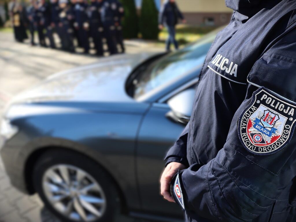 Policjant w mundurze z emblematem stoi obok radiowozu na tle grupy funkcjonariuszy.