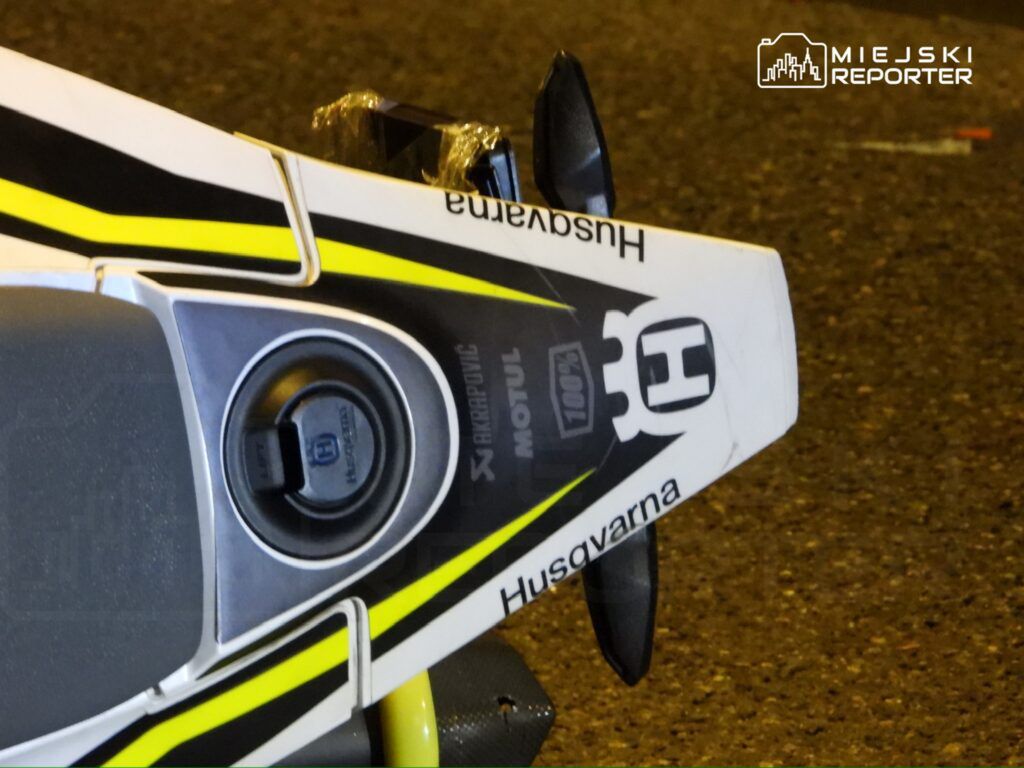 Motocykl marki Husqvarna z widocznymi detalami, takimi jak logo i elementy wykończeniowe, stoi na asfaltowej nawierzchni.