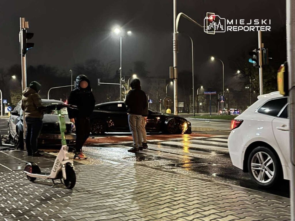 Grupa osób stoi przy skrzyżowaniu, obok zaparkowanego samochodu, w deszczową noc. Na chodniku widoczna hulajnoga elektryczna.