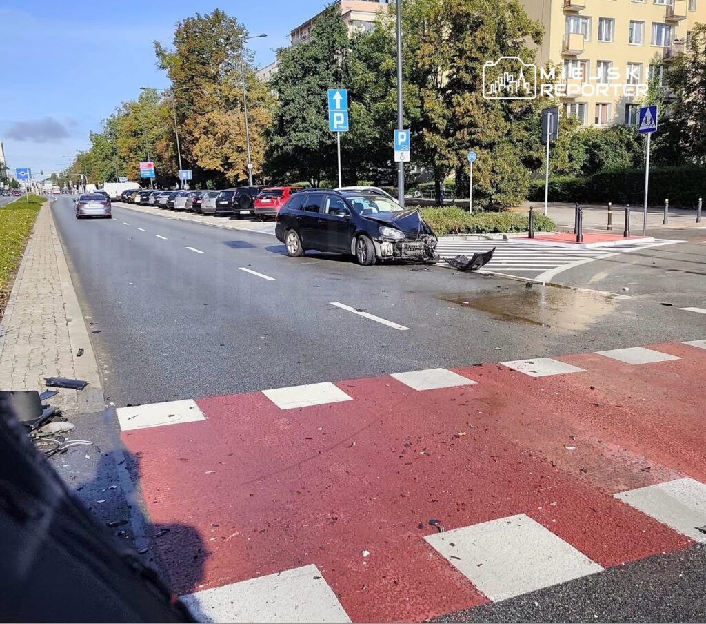 Zniszczony samochód osobowy z uszkodzoną przednią częścią stoi na jezdni obok zaparkowanych aut.