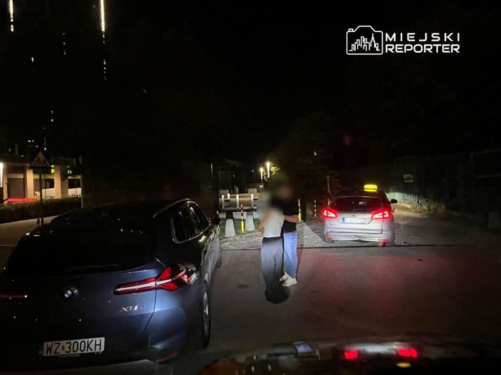 Mężczyzna i kobieta w ciemnym otoczeniu rozmawiają obok dwóch zaparkowanych samochodów.