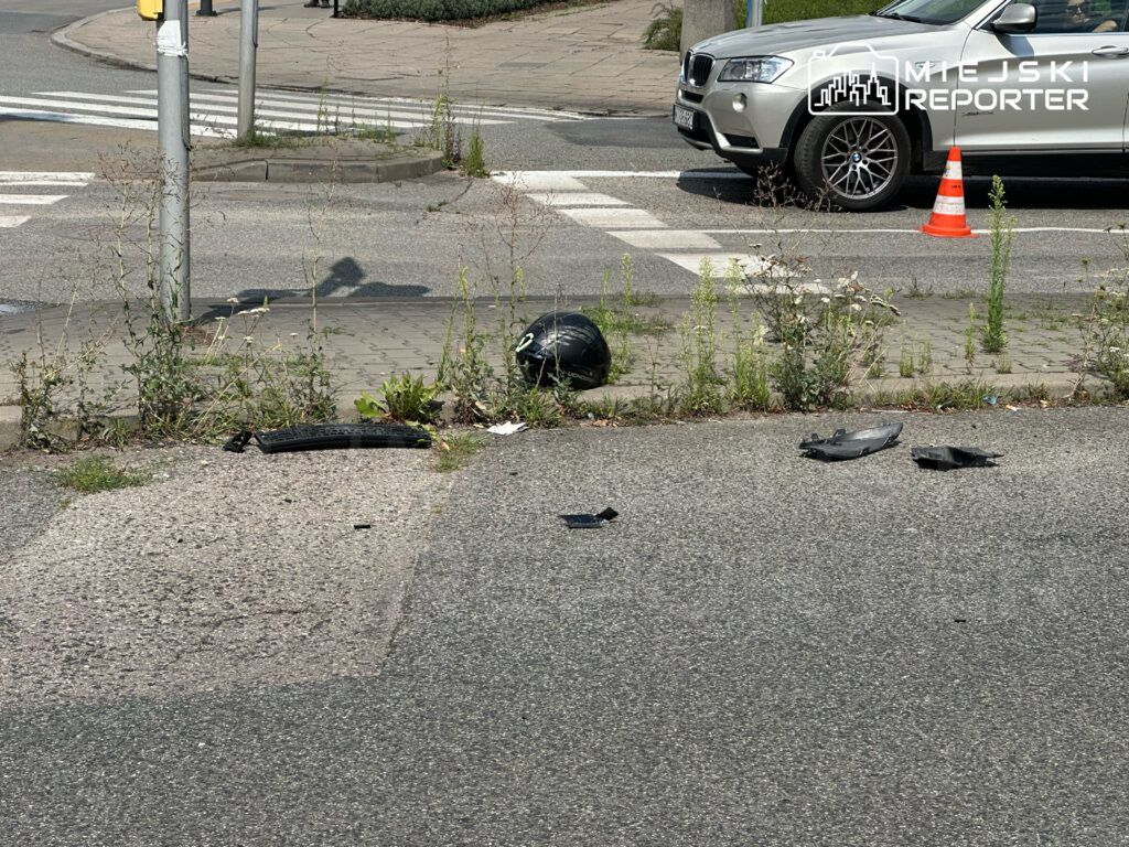 Zdarzenie drogowe przedstawiające uszkodzone elementy pojazdu oraz kask motocyklowy leżący na poboczu. W tle widoczny samochód osobowy oraz oznaczenie drogi. Miejsce wygląda na opuszczone, z chwastami rosnącymi wzdłuż krawędzi chodnika. Na jezdni brak innych pojazdów.