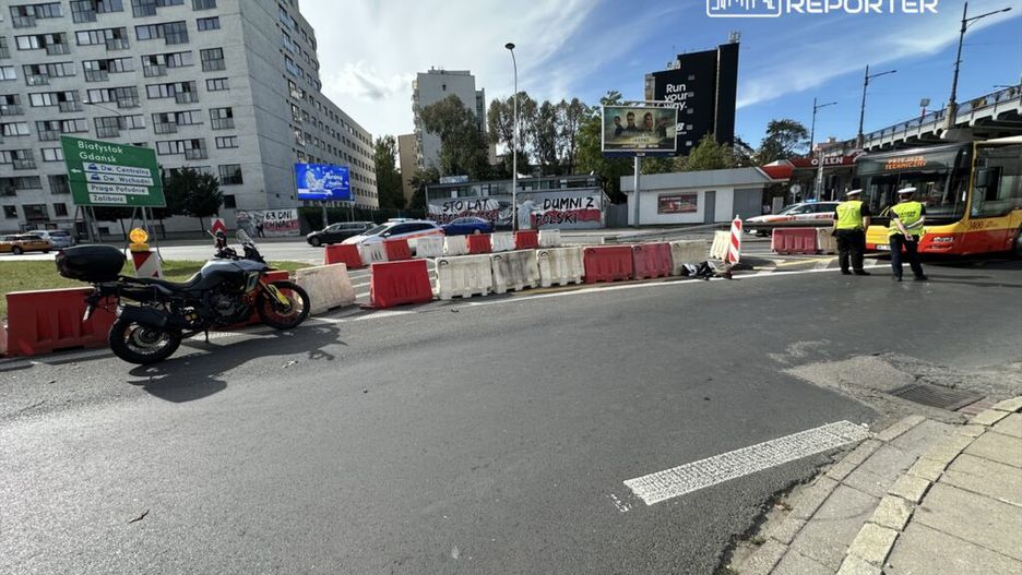 Motocykl stoi obok barier ochronnych na ruchliwej ulicy, a policjanci kontrolują ruch przy przystanku autobusowym.