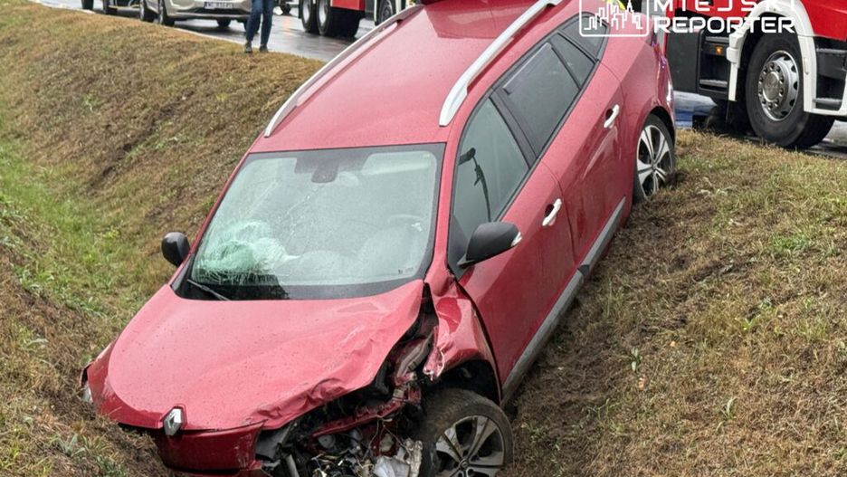 Czerwony samochód osobowy w rowie, z uszkodzoną przednią częścią, obok stojące pojazdy i wzywający pomoc strażak.