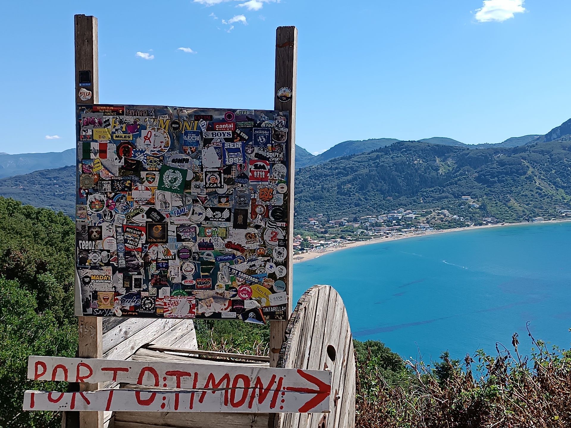 Drewniany drogowskaz z czerwonym napisem Porto Timoni i strzałką; w tle zatoka oraz plaża Agios Georgios Pagon na Korfu.