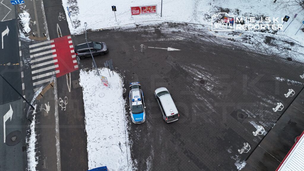 Policyjny radiowóz zaparkowany na skrzyżowaniu, obok przejścia dla pieszych, w otoczeniu zasypanej śniegiem nawierzchni.