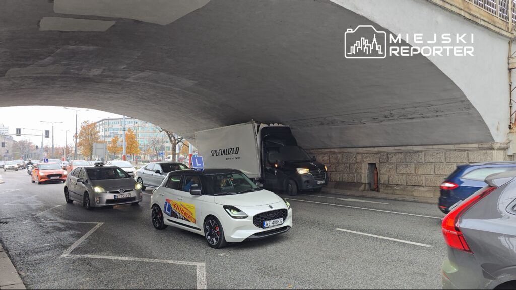 Ruch samochodowy pod wiaduktem, z ciężarówką utknętą w wąskim miejscu, w tle nowoczesne budynki i drzewa.