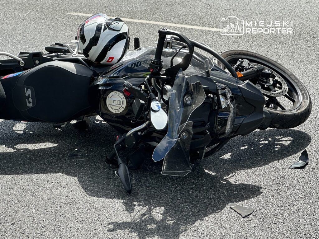 Motocykl leży na drodze, obok niego znajduje się kask. Na nawierzchni leżą fragmenty plastiku.