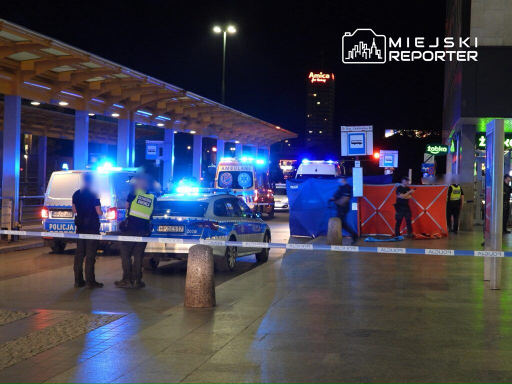 Policjanci w mundurach rozmawiają przed oznakowanym radiowozem, w tle karetka i niebieskie światła.
