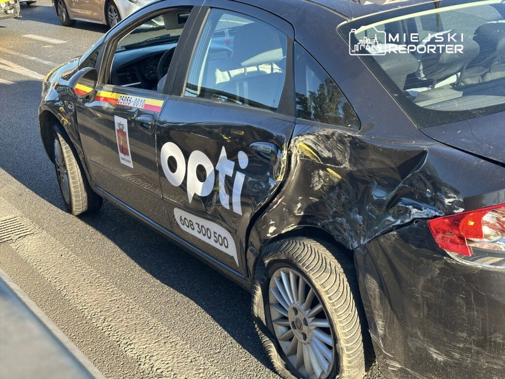 Uszkodzony czarny samochód osobowy marki opti z wgniecioną tylną częścią, zaparkowany na ulicy.