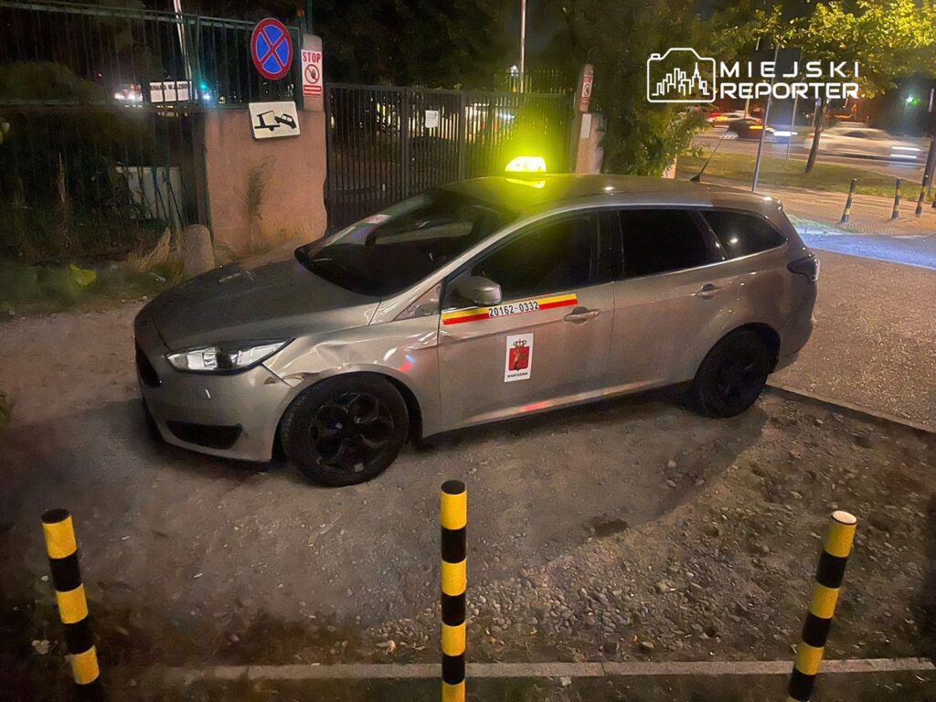 Samochód osobowy w kolorze srebrnym zaparkowany na chodniku, obok znaków zakazu parkowania oraz w pobliżu ogrodzenia.