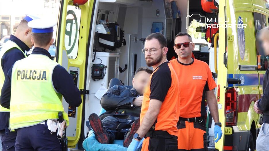 Ratownicy medyczni transportują poszkodowanego na noszach obok karetki, a policjanci zabezpieczają miejsce zdarzenia.