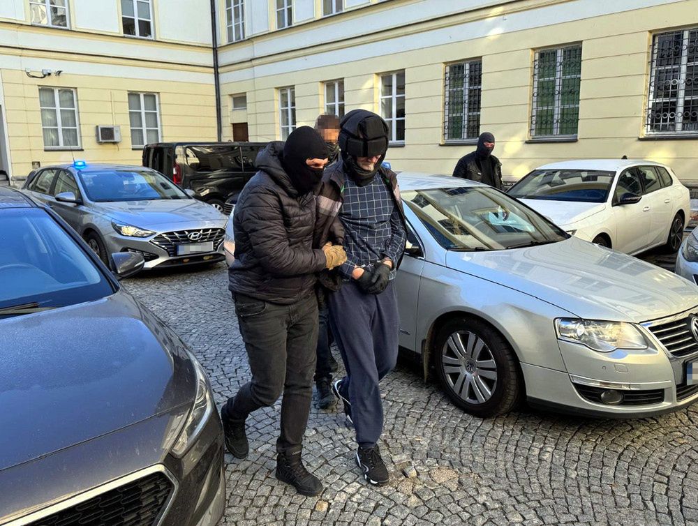 Mężczyzna w dresie prowadzony przez funkcjonariusza policji na parkingu między zaparkowanymi samochodami.