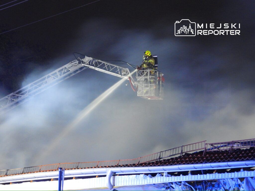 Strażak w podnośniku gaśniczym polewa wodą palący się budynek w gęstej mgle dymu nocą.
