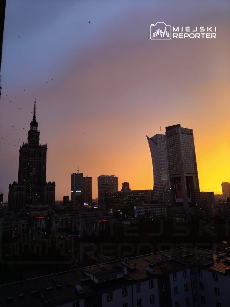 Warszawski krajobraz z dominującą wieżą z zegarem i nowoczesnymi budynkami, oświetlony ciepłym zachodem słońca.