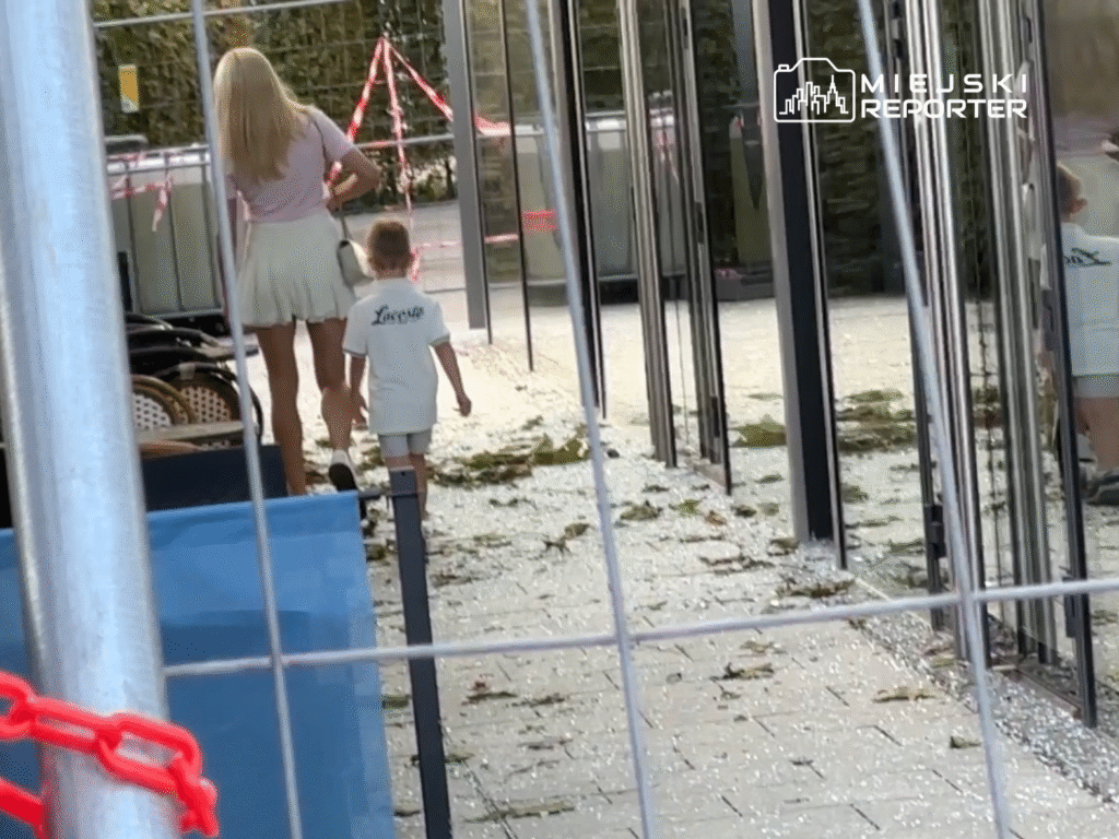 Kobieta w jasnej spódnicy idzie z dzieckiem w białym t-shircie wzdłuż ogrodzenia budowy, obok leżące liście.