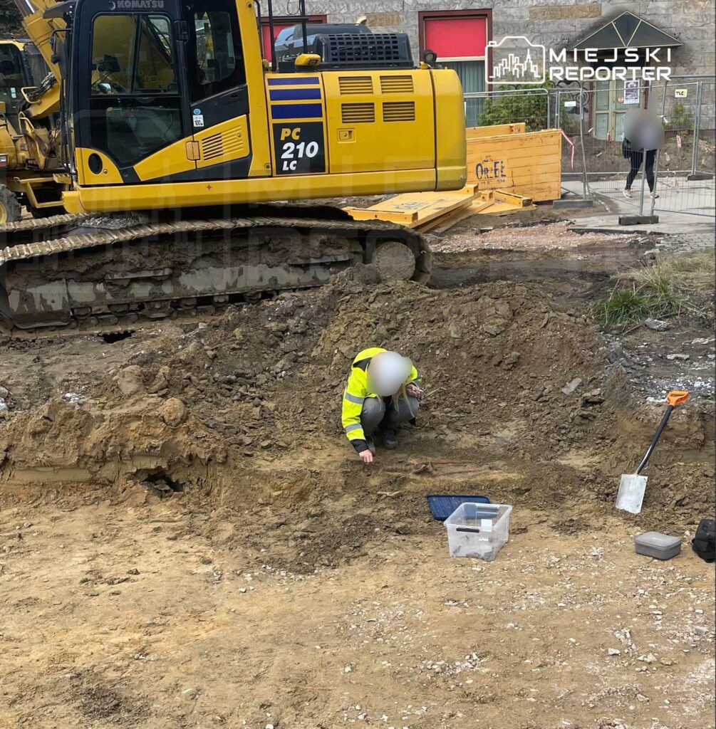 Pracownik w żółtej kurtce roboczej bada ziemię na placu budowy obok ciężkiego sprzętu.