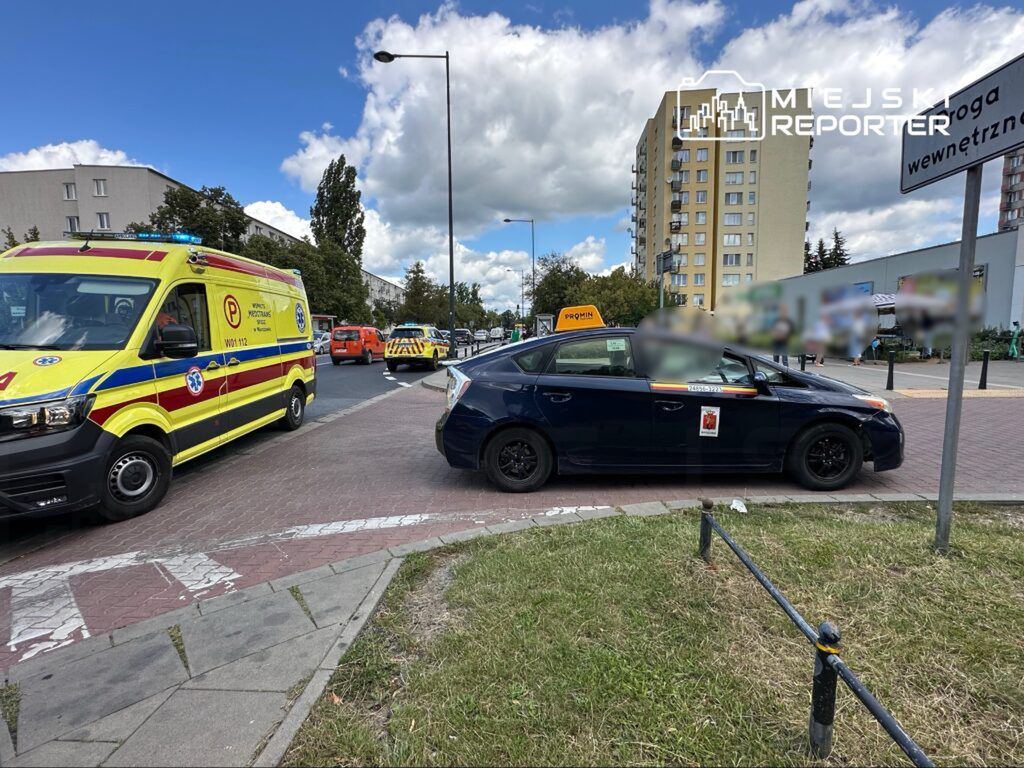 Karetka pogotowia i kilka innych pojazdów służbowych zatrzymanych na skrzyżowaniu, obok taksówki na chodniku.