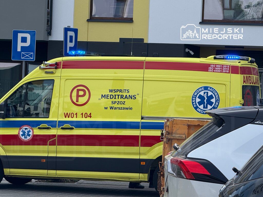 Zespół ratownictwa medycznego w żółtej karetce z niebieskimi światłami zaparkowany na ulicy.
