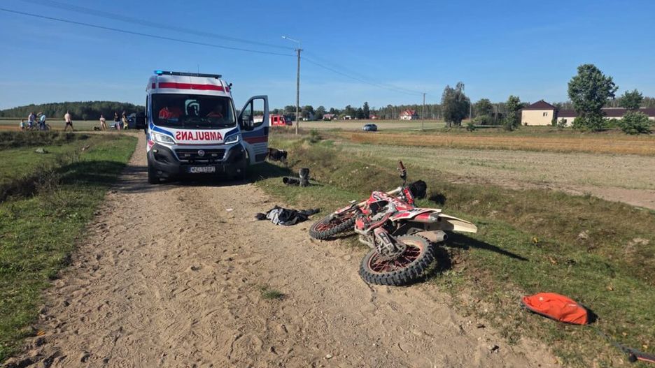 Ambulans z otwartymi drzwiami stoi na piaszczystej drodze obok przewróconego motocykla, a w tle widać grupę ludzi.