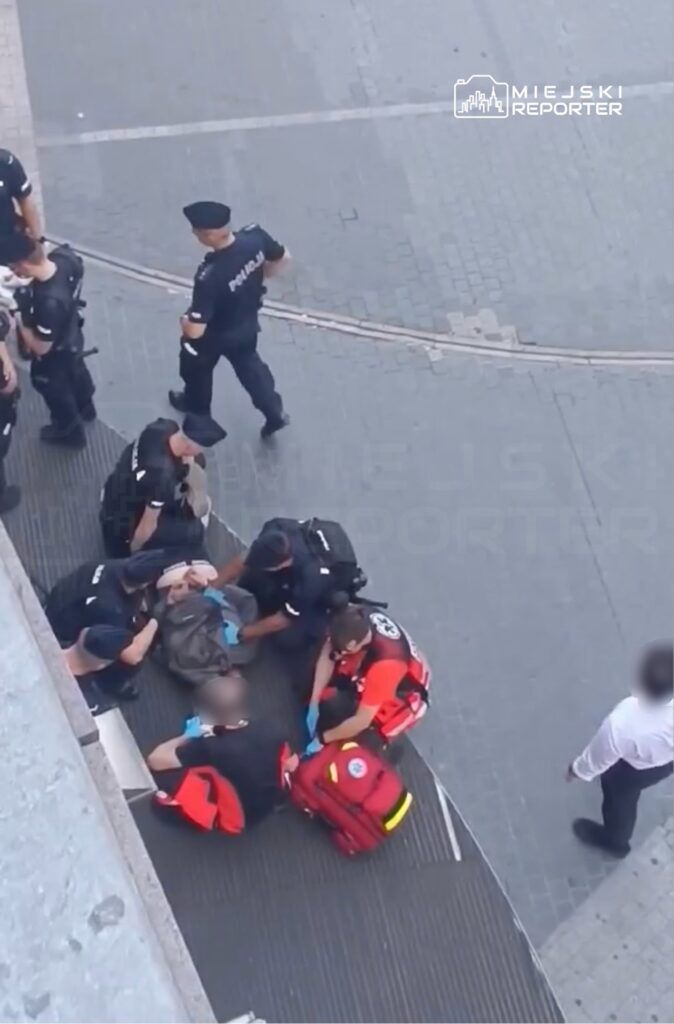 Grupa policjantów oraz ratowników medycznych udziela pomocy mężczyźnie leżącemu na chodniku w miejskim otoczeniu.