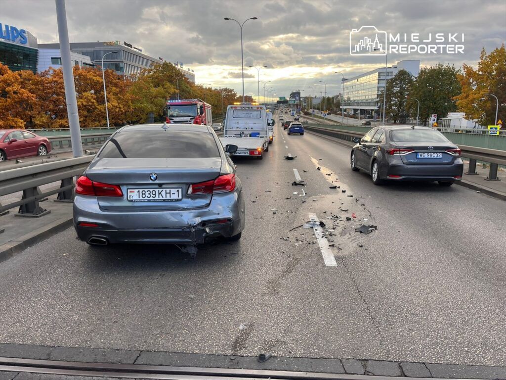Uszkodzony samochód osobowy stoi na jezdni, obok leżą resztki zderzenia, w tle inne pojazdy i biurowce.