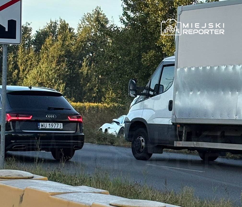 Czarny samochód osobowy zatrzymany na drodze obok białego pojazdu dostawczego, otoczone drzewami.