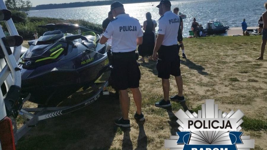 Dwóch policjantów w mundurach rozmawia obok skuteru wodnego na plaży nad jeziorem. W tle widać ludzi wypoczywających nad wodą.