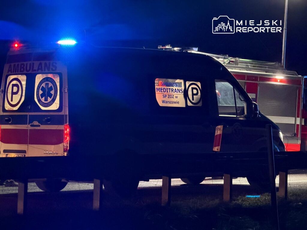 Ambulans z niebieskimi światłami zaparkowany na drodze, z widocznym znakiem "MEDITRANS" oraz informacją o strefie parkingowej.