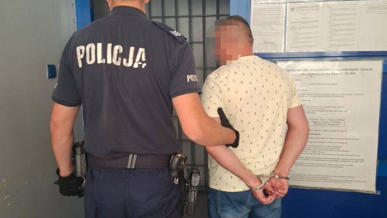 Policjant w mundurze prowadzi zatrzymanego mężczyznę z rękami związanymi z tyłu w korytarzu komisariatu.