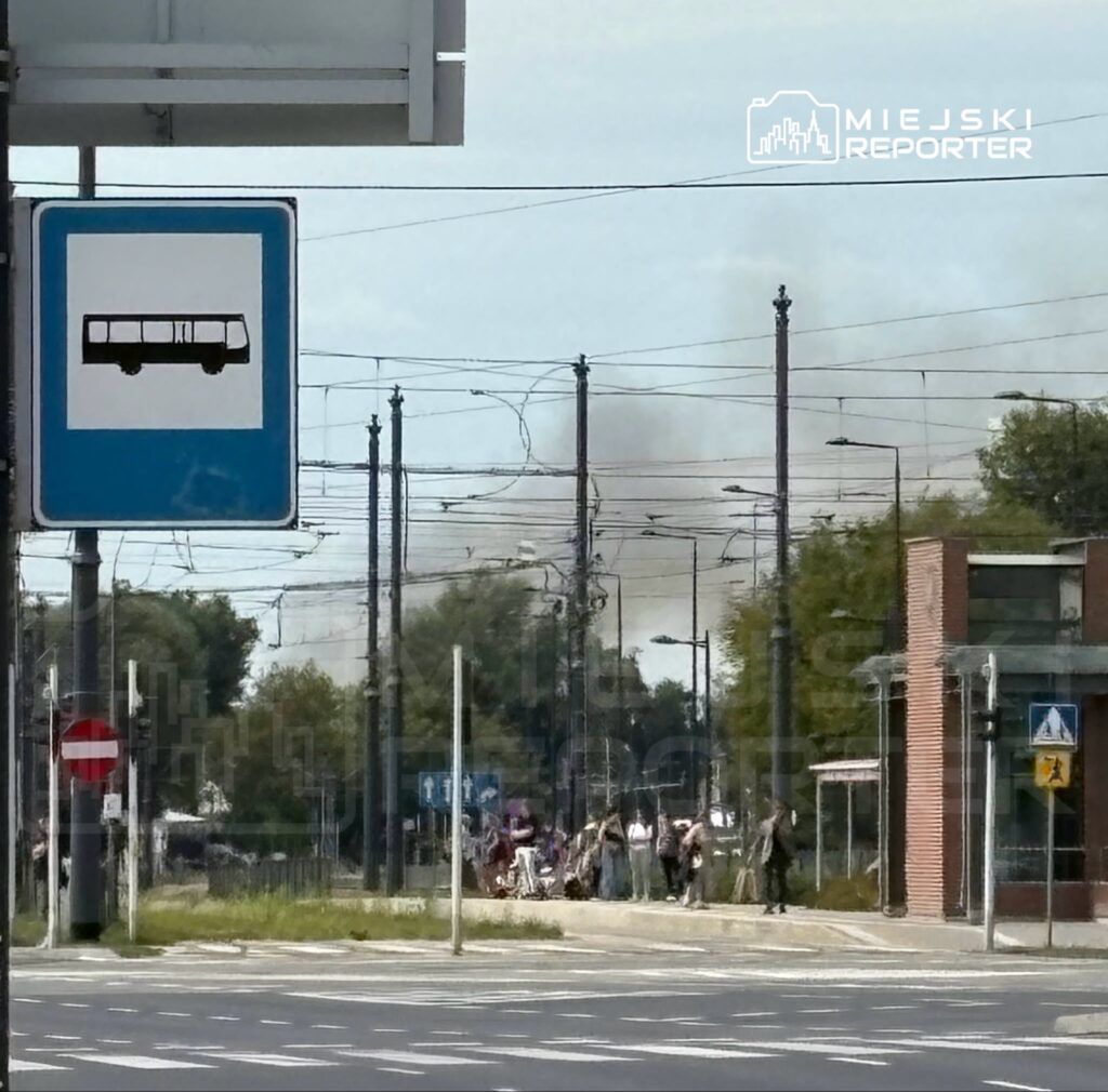 Grupa osób czekająca na przystanku tramwajowym, w tle widoczny dym unoszący się z kierunku torów.