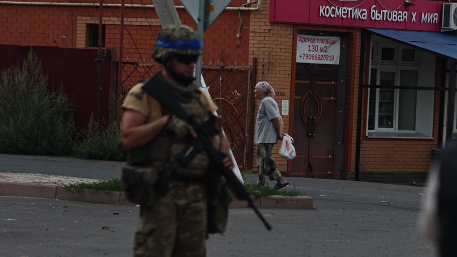 SUDZHA, RUSSIA - AUGUST 16: A Ukrainian military member holds a weapon, and a woman walks a street on August 16, 2024 in Sudzha, Russia. The fighting in the Kursk Oblast began on August 6, 2024, when the Armed Forces of Ukraine crossed the Russian-Ukrainian border near the city of Sudzha and began to advance deep into Russian territory, and in a few days took control of dozens of settlements in Kursk region. (Photo by Yan Dobronosov/Global Images Ukraine via Getty Images)