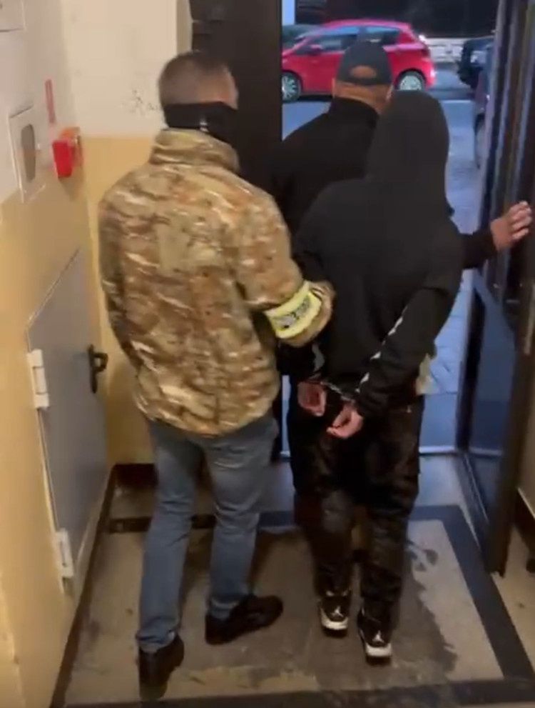 Mężczyzna w mundurze trzyma rękę skuta w kajdankach młodego mężczyzny, wychodząc z budynku.