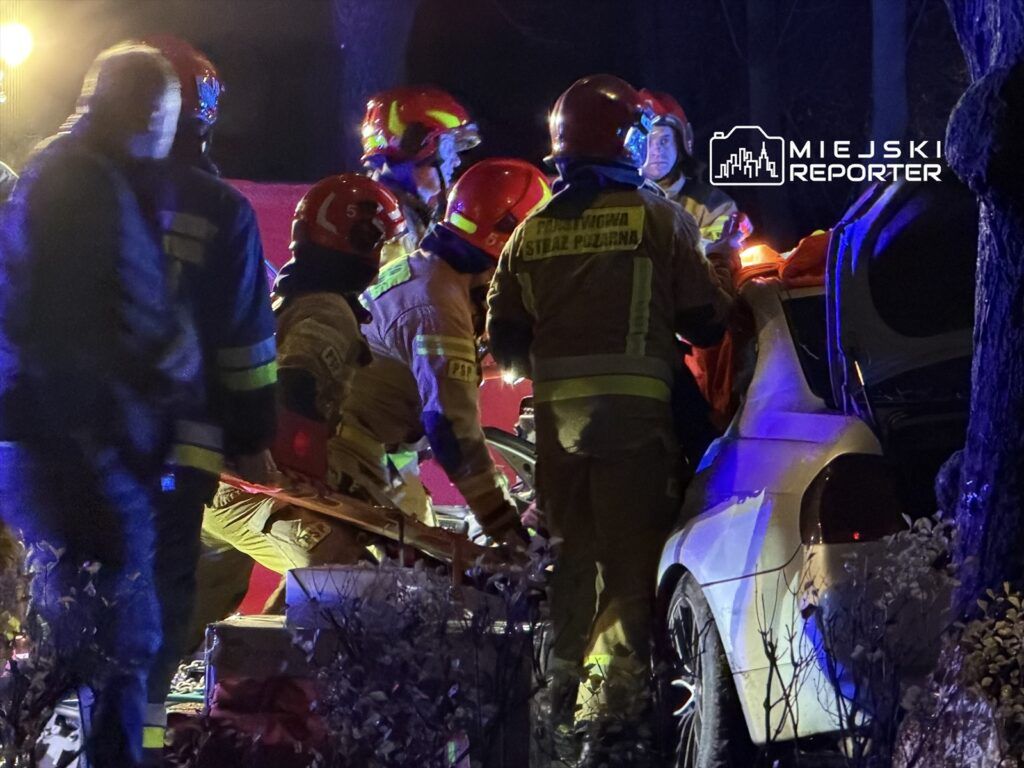 Grupa strażaków w hełmach udziela pomocy poszkodowanemu przy uszkodzonym samochodzie w nocy.